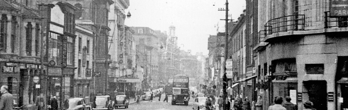 A black and white photograph of Granby Street, Leicester. There are people walking on the pavement and cars parked along the street. In the middle of the road in the foreground is a Keep Left bollard.