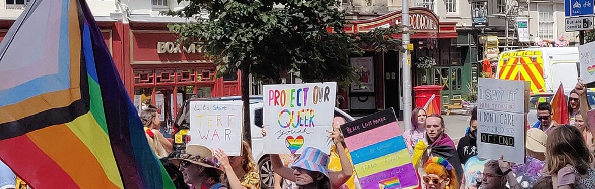 Bristol Pride Parade 2022: a group of people hold rainbow flags and signs as they march past the Bristol Hippodrome