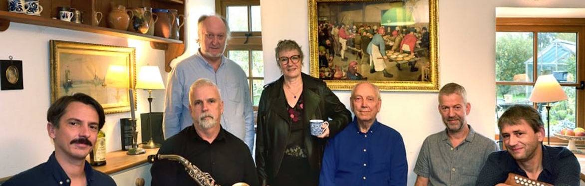 7 members of the band: 5 sitting in a row, 2 standing behind. Some are holding instruments - a saxophone and a diatonic accordian. Behind them are a selection of paintings on the walls.