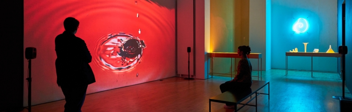 Installation view of the exhibition Haunted Ecologies, with a large red film projectuion of water dripping and two people watching the film.