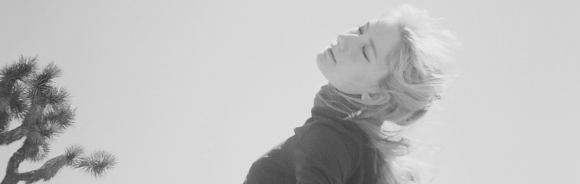 Black-and-white photo of artist Hannah Herschend standing outdoors in a desert landscape, with Joshua trees visible in the background. She leans backward with eyes closed and arms extended behind her,