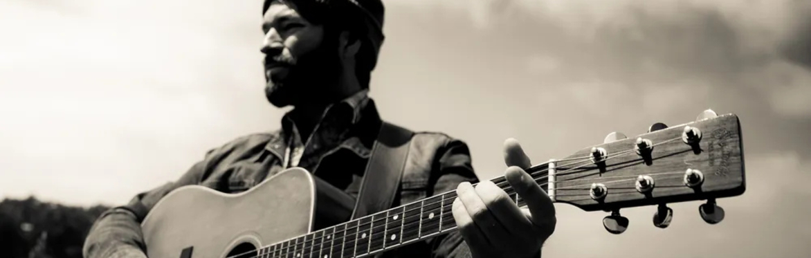 Black-and-white promotional photo of Joel Van Horne of Covenhoven standing outdoors in a grassy field with an acoustic guitar, wearing a knit cap and looking toward the horizon beneath a cloudy sky.