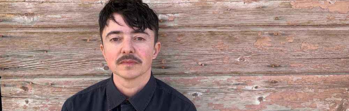 Dean Spade, a white trans person with short straight dark brown hair and a mustache stands in front of a wooden wall. He wears a dark grey button up shirt.