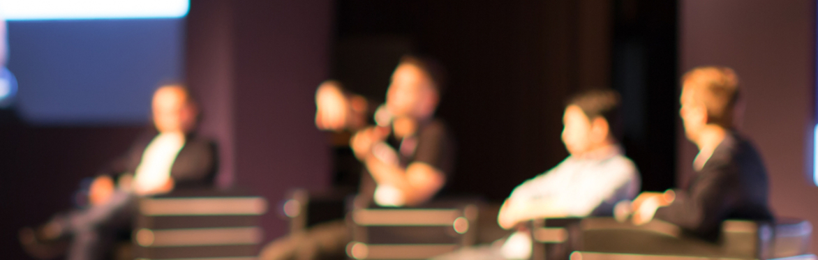 Four adults sat on chairs on a stage. One adult has a microphone in his hand.