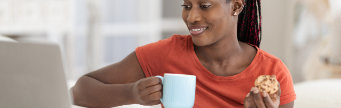 Woman looking at a computer while smiling and holding a cup of coffee and muffin