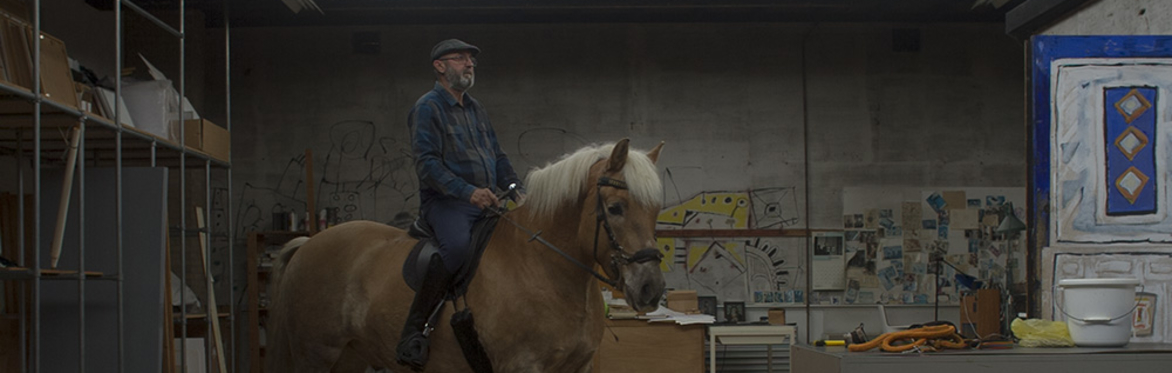 A lightskinned man with a grey beard is sat on a brown horse in an artists' studio