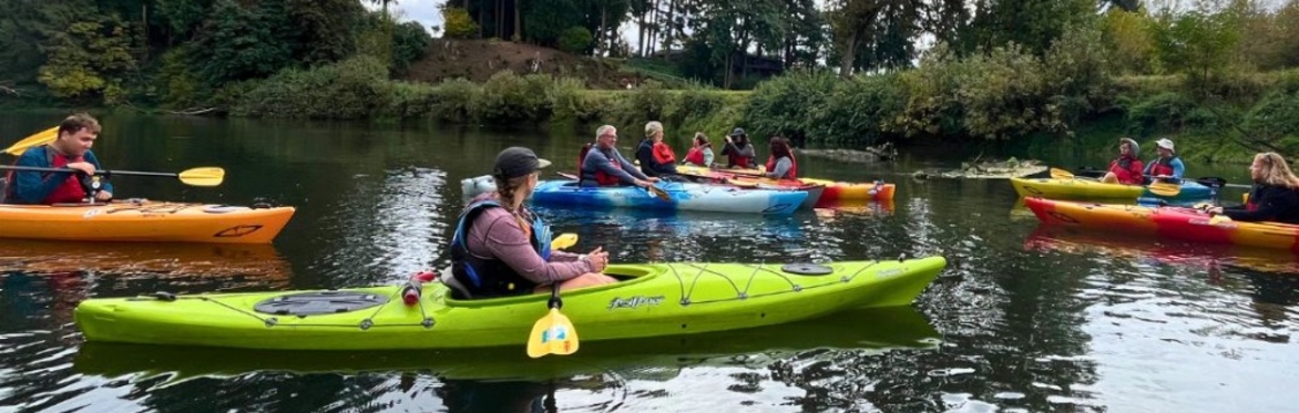 A group of people in single and tandem (2 person) kayaks wearing P-F-Ds (lifejackets) float on calm water