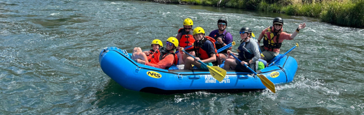 A group of paddlers on a bright blue raft on clear water on a sunny day smile at the camera while wearing P-F-Ds (lifejackets) and helmets. The raft guide waves to the camera.