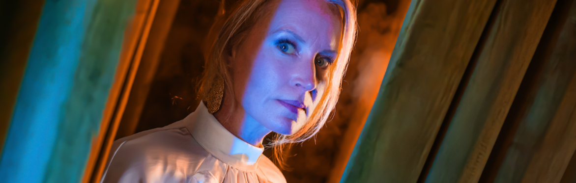 A white woman in a white blouse stares at the camera through a pair of green curtains. Orange light bathes her face and catches some haze in the background.