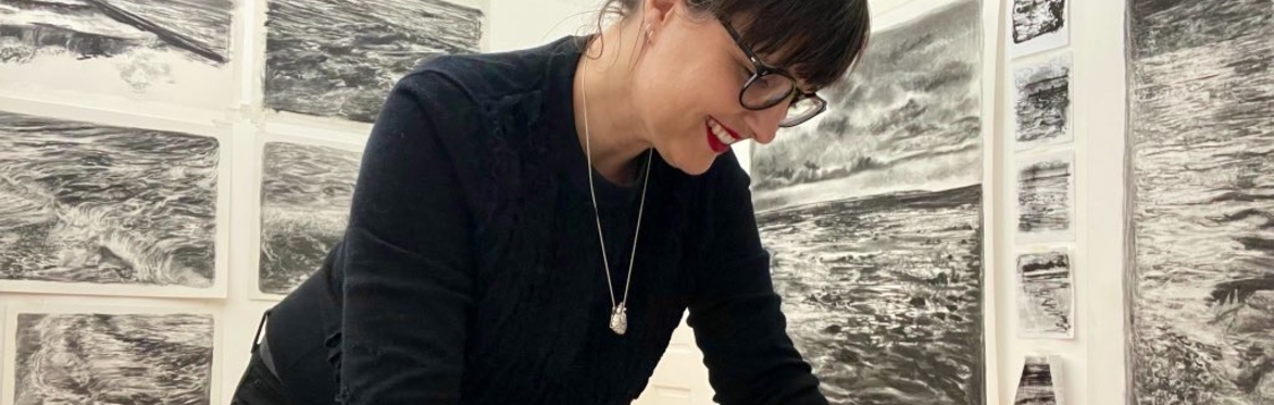 Liz Atkin drawing with charcoal in her studio with large charcoal drawings behind her