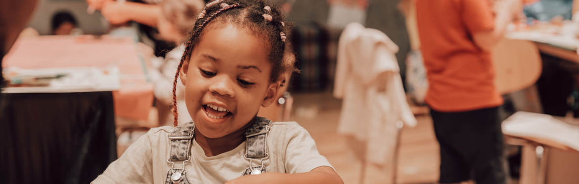 A young child sits at a table cutting paper, other families can be seen in the distance making together.