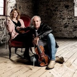 The image features a man wearing blue jean and white tshirt and black jacket sitting on the floor, sittingbehind him on a chair is a woman in a light pink cardigan and black top holding fiddles