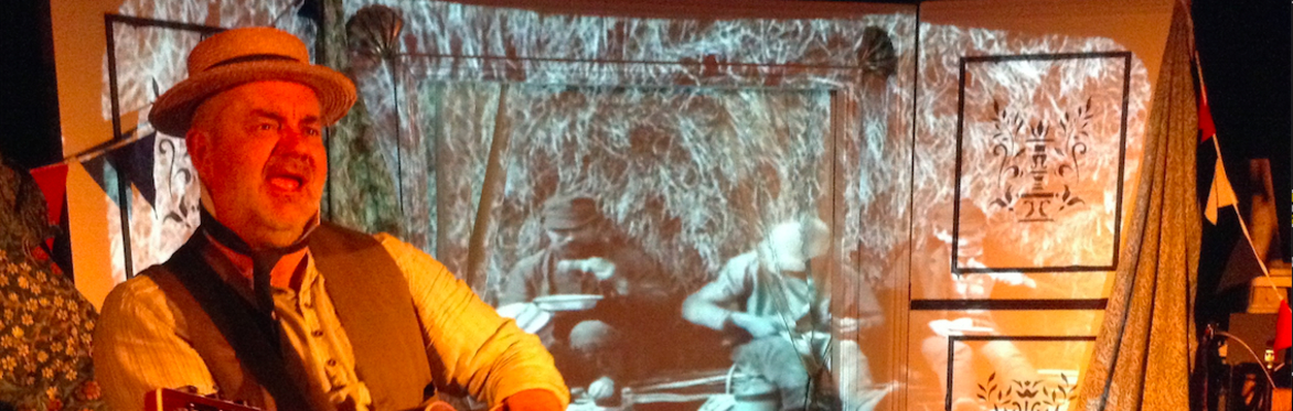 Man with a boater hat, singing with a guitar against a backdrop of a picture of working men from the 1900's sitting on the floor and eating