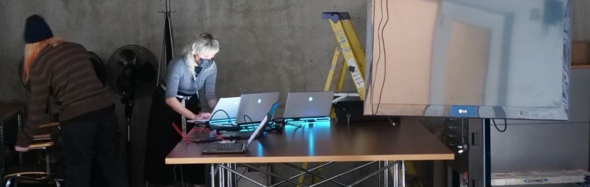 2 people surrounded by glowing laptops, a TV and various cables.