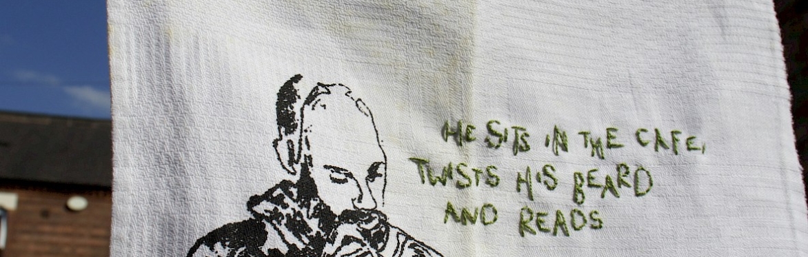 An embroidered handkerchief showing a person sat in a cafe reading a book. The words 'He sits in the cafe, twists his beard and reads' is stitched in the centre.