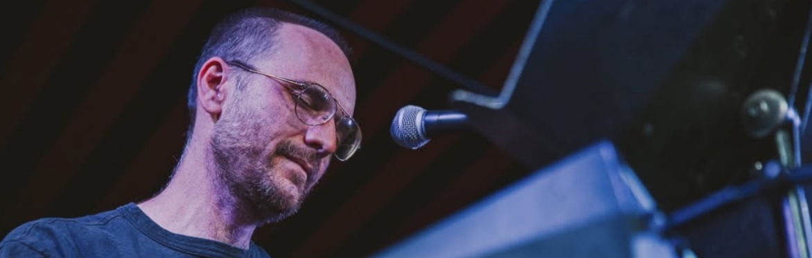 Musician Ben Vida performs on stage, focused intently while playing a keyboard. He wears round glasses and a dark t-shirt, lit in cool tones of blue and purple from stage lights. A microphone is posit