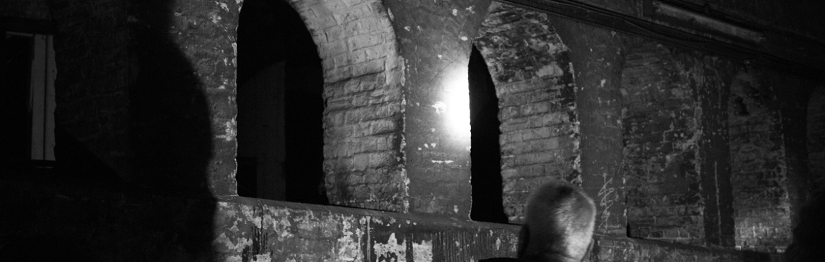 Black-and-white photo of a dim underground brick passage. Three arched openings line the old wall, with a small exposed lightbulb casting a stark glow.