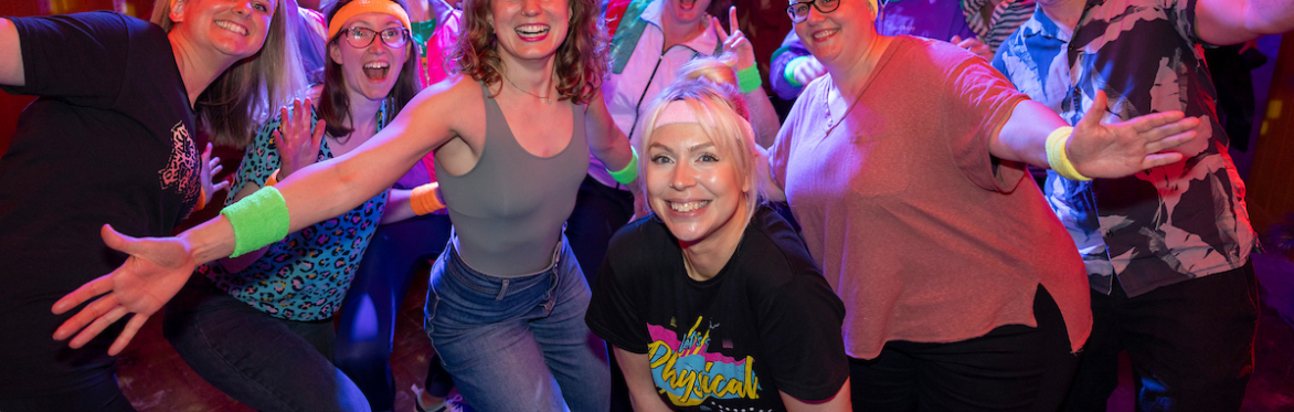 a group of smiling improvisers with audience behind cheering, dressed in 1980s fitness costumes