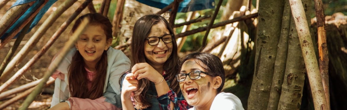 Three girls laughing in a home made den of sticks with dirty faces.