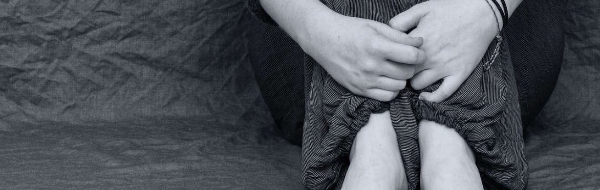 someone, possibly a teenager, is sat against a wall - knees pressed to the chest and arms around their shins