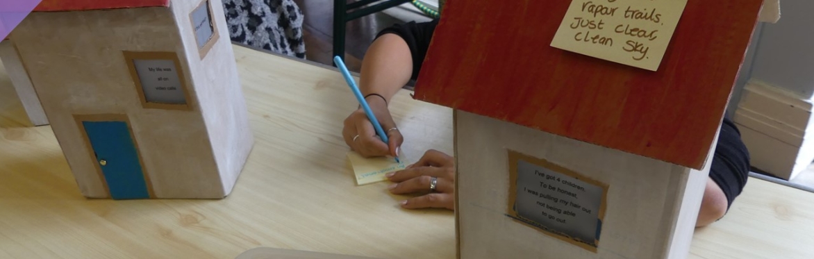 a woman's hands with bare arms up to the elbow, writing on a post-it note. Each side there are two 3-d models of houses, each about 30 cm high with red rooves and white walls