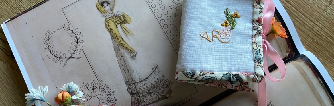 Handmade embroidered needle cases on an open book of Regency fashion illustrations, one open with needles in blue felt, tied with pink ribbon.