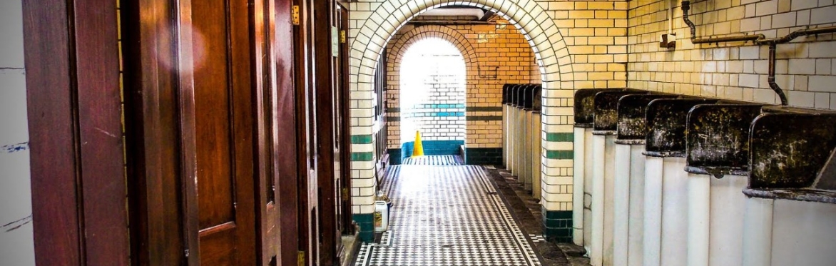 The interior of a men's public toilet. On the right there is a row of urinals in waist height booths, covered in white tiles. ON the left at the wooden doors to WCs. The floor between is of black and
