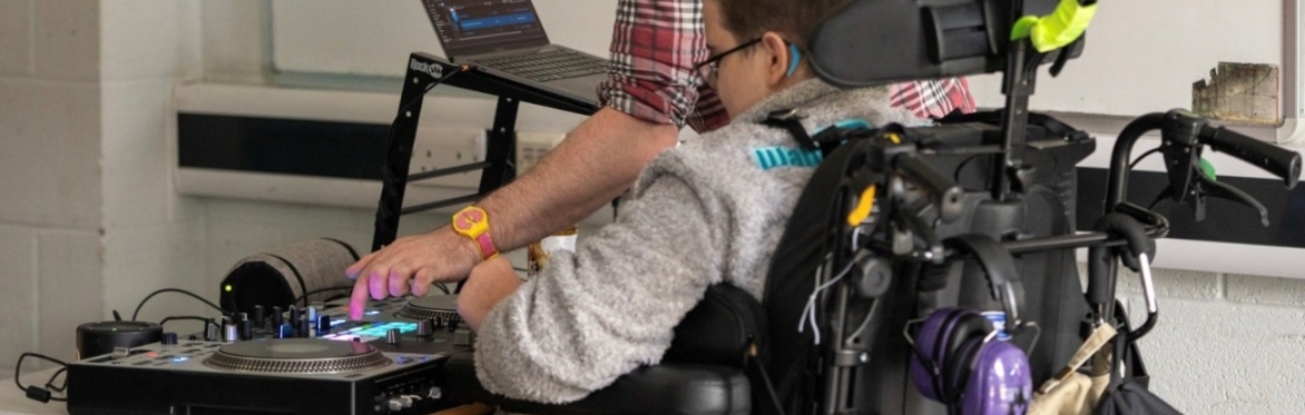 A child in a wheelchair with SEND is exploring and playing with a DJ deck, assisted by an adult who is standing beside them.