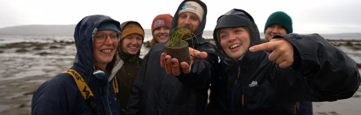 6 smiling people standing on a beach in waterproofs and hats. The one in the middle is holding a core of sediment and seagrass.