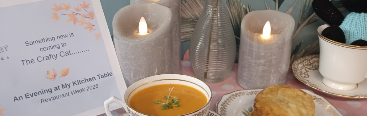 An arrangement featuring soup in a vintage tea cup, a mini cheese scone on a china plate, with candles, a little notice, and a knitted cat in a tea cup, against a background of floral wallpaper.