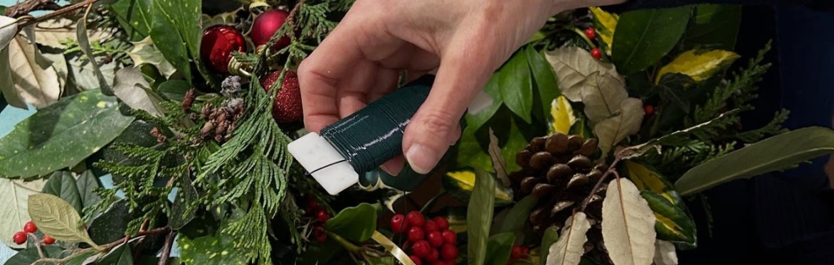 a hand is winding green wire around a wreath made of green leaves, with red berries and pinecones