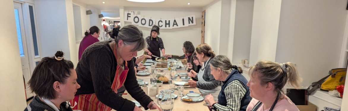 A group of people wearing aprons sit around a long rectangular wooden table and are busy slicing bread and eating soup. At the end of the room