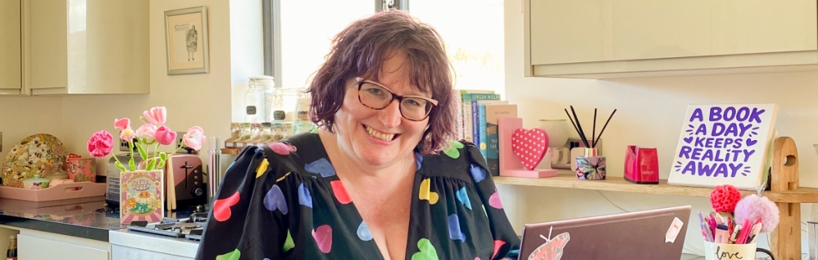 Claire dressed in a pretty black dress with colourful heart print on it sitting in her kitchen in fron of her laptop on the worktop and a sign that says a book a day keep reality away