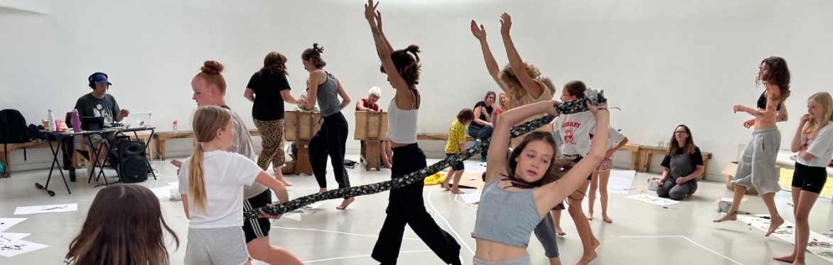A picture of people dancing and playing with fabrics. There is someone in the background playing music and a child in the foreground wearing many net skirts