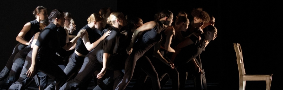 The image depicts a group of dancers in synchronized motion on a dark stage, all dressed in black attire. They are leaning forward, intensely focused on a single empty wooden chair illuminated by a sp
