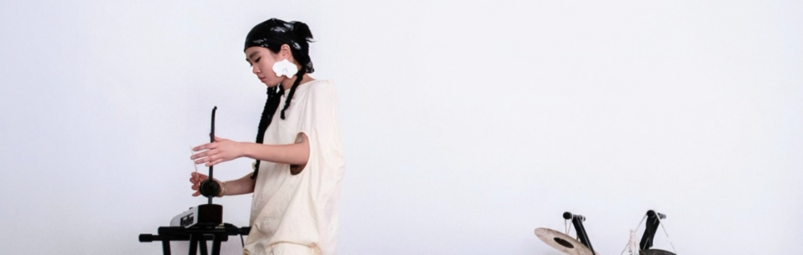 A woman stands in front of a white wall playing a theremin