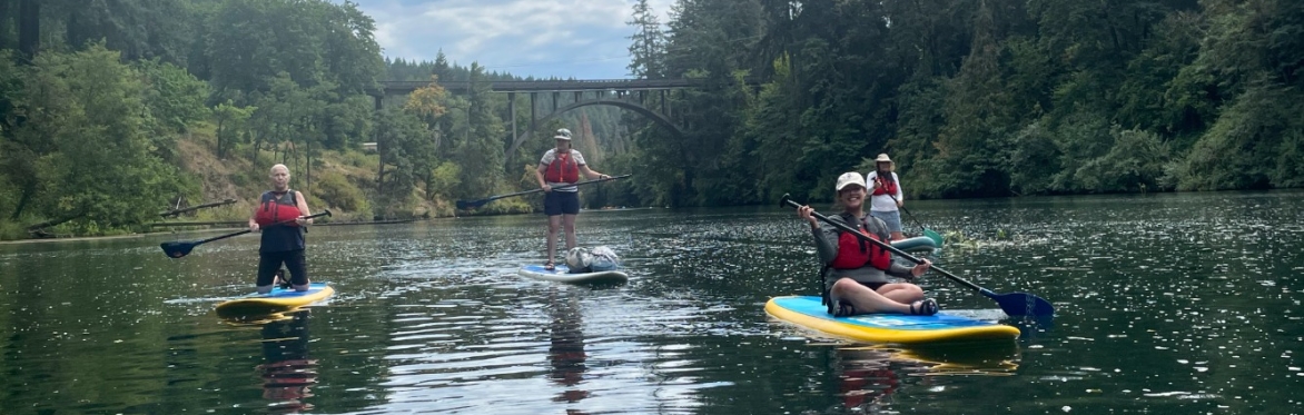 4 people on paddleboards sit, kneel, or stand as they smile for the camera. Everyone is wearing a P-F-D (lifejacket)