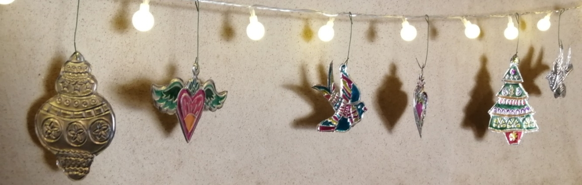 A String of white fairy lights with Mexican colourful tin bauble, heart with wings, bird, Christmas tree and star decorations hanging, casting a shadow on the wall behind..