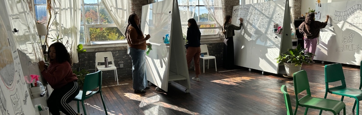 People are working in a sunny studio with hardwood floors, drawing on whiteboards.