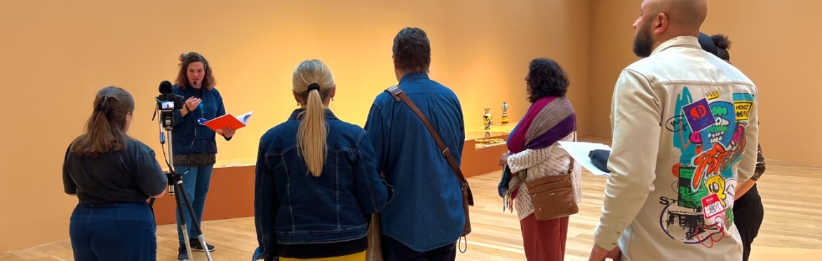Group of people in a gallery space, listening to a speaker. There is a camera filming the speaker and a display of ceramics in the background.