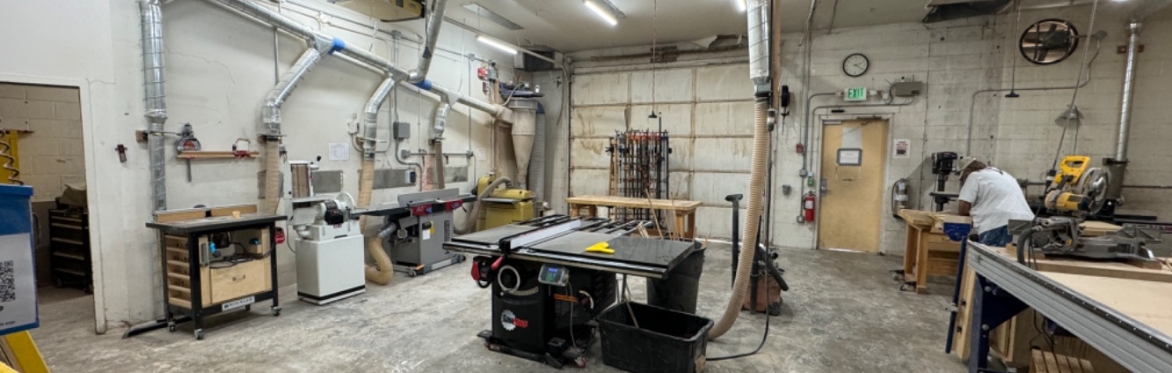This is a woodshop with floor power tools around the outside walls and a SawStop table saw in the center. There is a person working at one of the work benches.