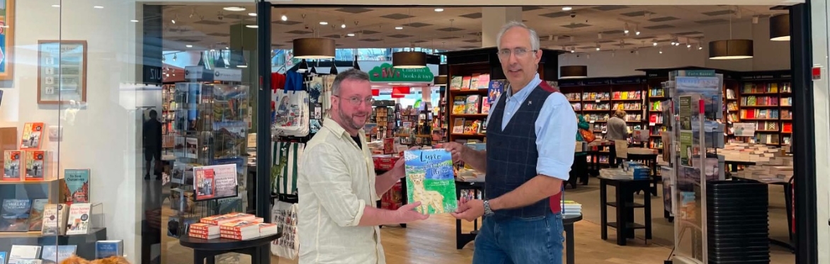Colour Pic of Sam wearing a blue shirt with a dark blue waistcoast and blue jeans handing a copy of this book about Alpacas to Waterstone's manager Toby outside of the Waterstones Inverness Shop.