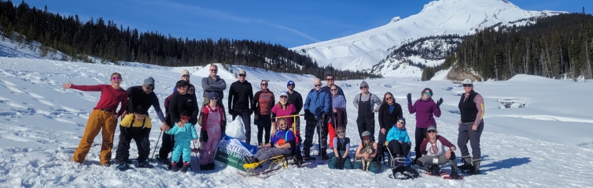 A large group of participants, staff, and volunteers pose together in front of snowy Mt Hood on an inclusive and adaptive snowshoe trip