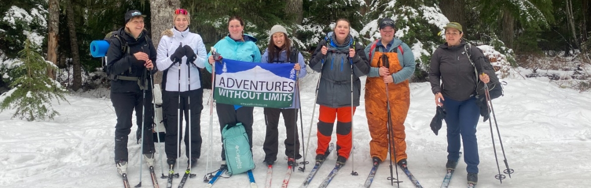 7 people wearing snow gear and cross country skis pose with the A-W-L flag on a snowy trail