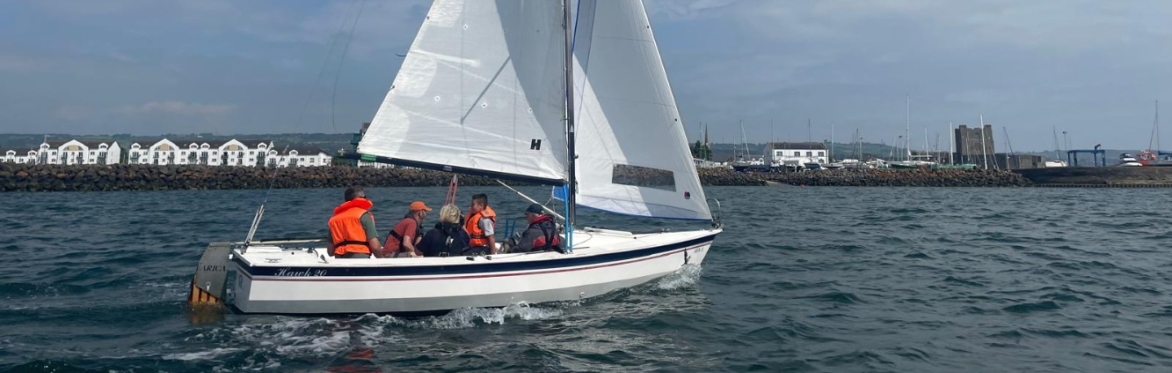 A small white sailing boat with raised sails carries a group of people wearing orange life jackets across water