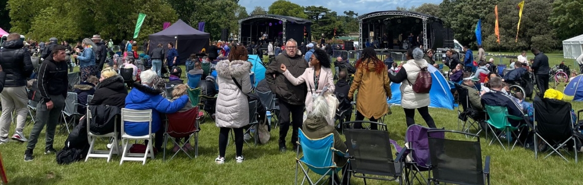 Large outdoor music festival, showing two large stages with flags on either side.  Some people sitting in chairs facing the stages, some dancing around and looking happy. Photo taken at 2024 event
