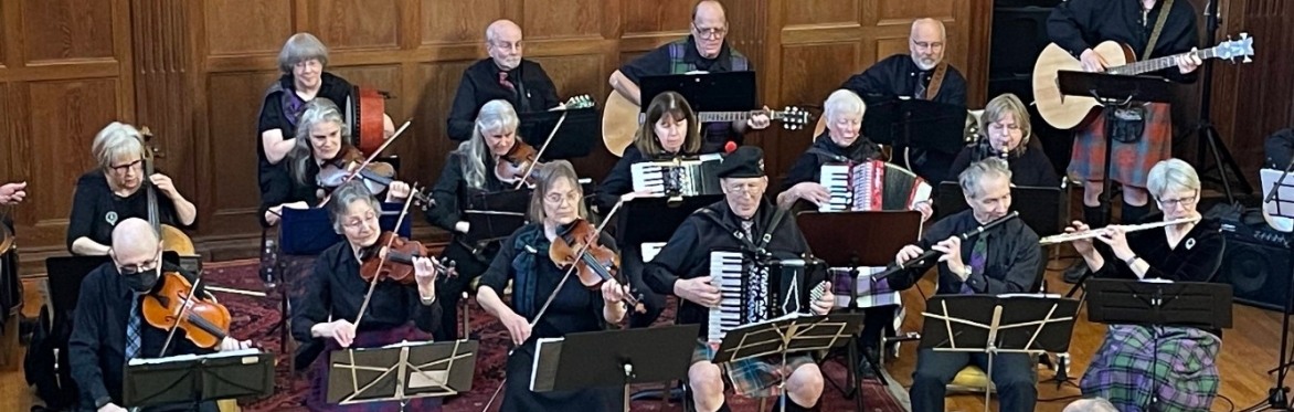 A group of orchestra musicians playing accordion, violin, guitar, wind instruments and percussion. They are seated together with music stands in a room with wood paneling.
