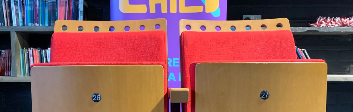 Two wooden theatre seats with red fabric in front of a Middle Child banner.
