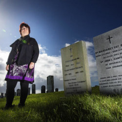 Fiona dressed in dark jacket and legging with a flowery purple dress, stood next to the white gravestone of Margaret Fay Shaw with a blue sky in the background and green grass in the foreground
