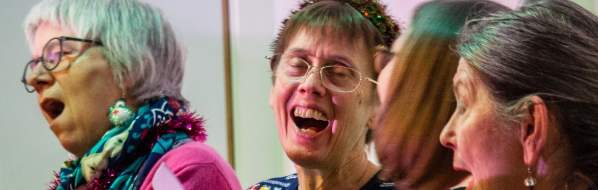 Three older women at a festive gathering; the woman on the left, wearing glasses and a Christmas-themed sweater, sings heartily while the other two women face toward her.